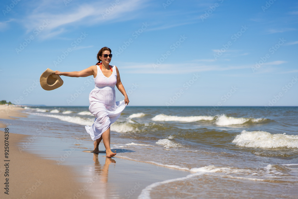 Summer holidays. Young beautiful woman wearing orange bikini and long white dress and sunglasses running with beige hat in her hand on sandy beach on sunny day. Energy, joy of life and youth. Summer holidays. Young beautiful woman wearing orange bikini and long white dress and sunglasses running with beige hat in her hand on sandy beach on sunny day. Energy, joy of life and youth.