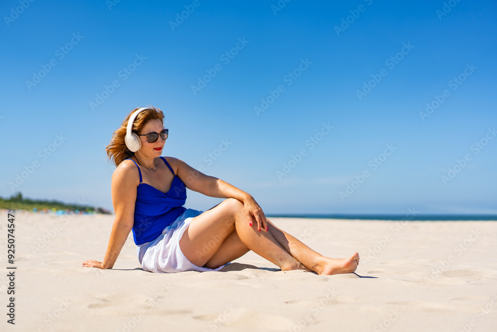 Summertime with music. Beautiful young woman in blue swimsuit and white sarong and sunglasses listening to music in headphones while sitting on white sandy beach on background of blue clear sky. Summertime with music. Beautiful young woman in blue swimsuit and white sarong and sunglasses listening to music in headphones while sitting on white sandy beach on background of blue clear sky.