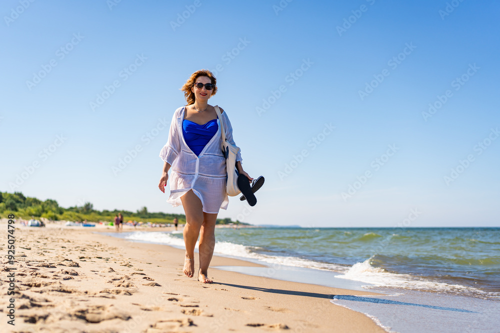 Summertime by Baltic Sea. Beautiful young woman in blue swimsuit and white beach dress and sunglasses with beach bag and tip toes in hand walking by sea on sandy beach on beautiful sunny day. Summertime by Baltic Sea. Beautiful young woman in blue swimsuit and white beach dress and sunglasses with beach bag and tip toes in hand walking by sea on sandy beach on beautiful sunny day.