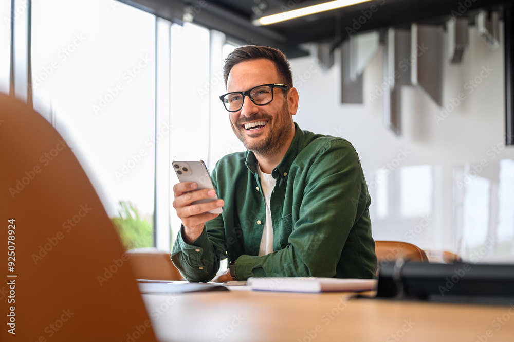 © Moon Safari - Cheerful handsome businessman in eyeglasses looking away while using mobile phone at desk in office