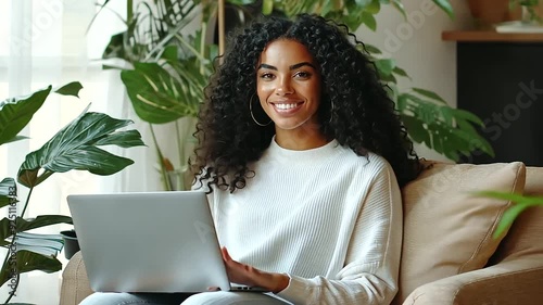 A beautiful young woman seated on a cozy armchair, smiling at the camera while holding a laptop on her lap. The image highlights a relaxed and productive workspace, ideal for portraying modern freelan photo