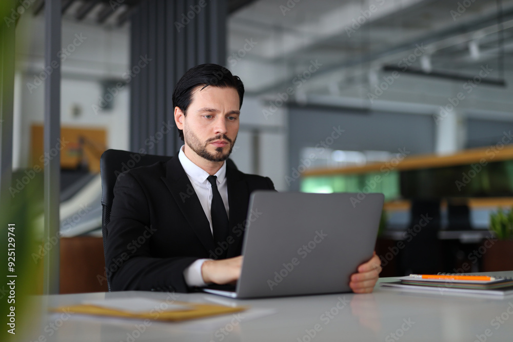 A man in a suit is sitting at a desk with a laptop open in front of him. He is focused on his work, possibly typing or browsing the internet. Concept of professionalism and productivity A man in a suit is sitting at a desk with a laptop open in front of him. He is focused on his work, possibly typing or browsing the internet. Concept of professionalism and productivity