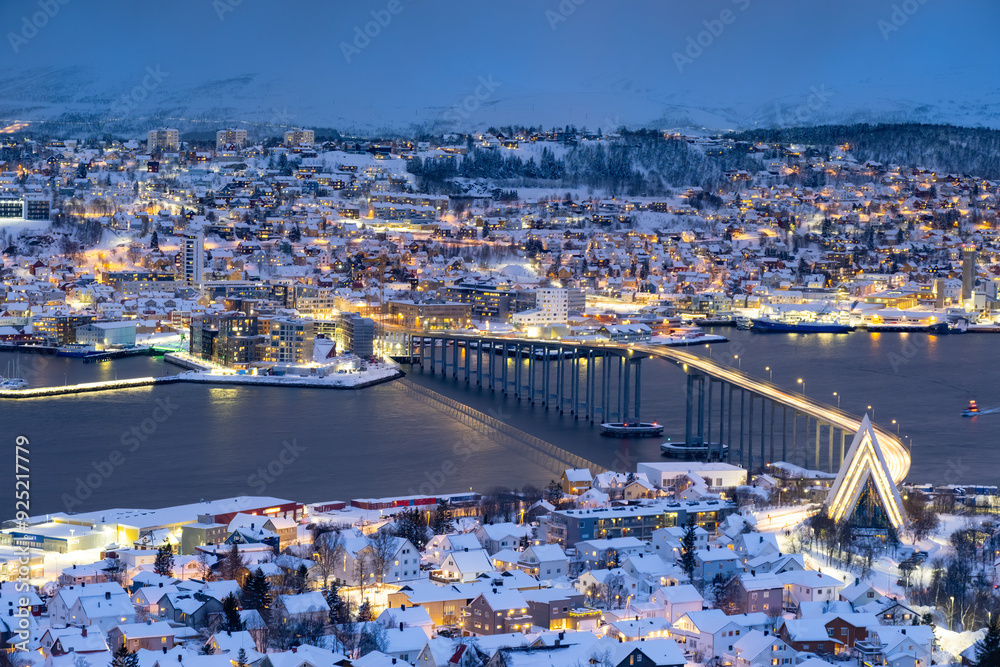 Obraz premium Panorama of Norwegian city of Troms in the winter. Snowy roofs, bridge, embankment near the port and fishing ships. Evening lights