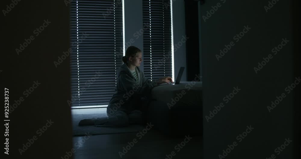 A woman uses a laptop while sitting on the floor near the bed at home. Closed blinds, dark room