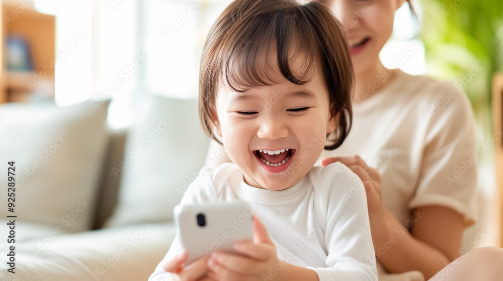 Toddler holding a smartphone laughs while sitting with their mother on a cozy couch in a bright living room Toddler holding a smartphone laughs while sitting with their mother on a cozy couch in a bright living room