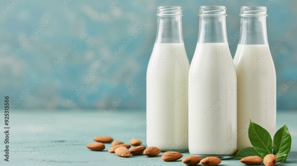 Three glass bottles of almond milk with almonds and green leaves on a wooden table against a blue background. Three glass bottles of almond milk with almonds and green leaves on a wooden table against a blue background.