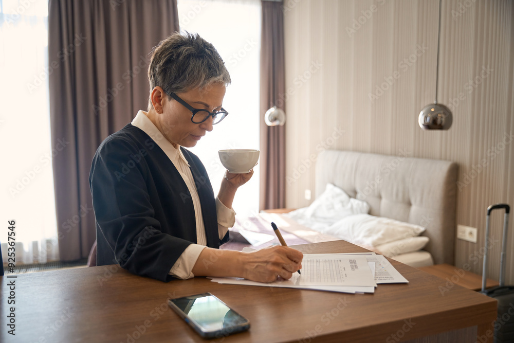 Senior woman filling out the form, holding cup of tea, standing in hotel room Senior woman filling out the form, holding cup of tea, standing in hotel room