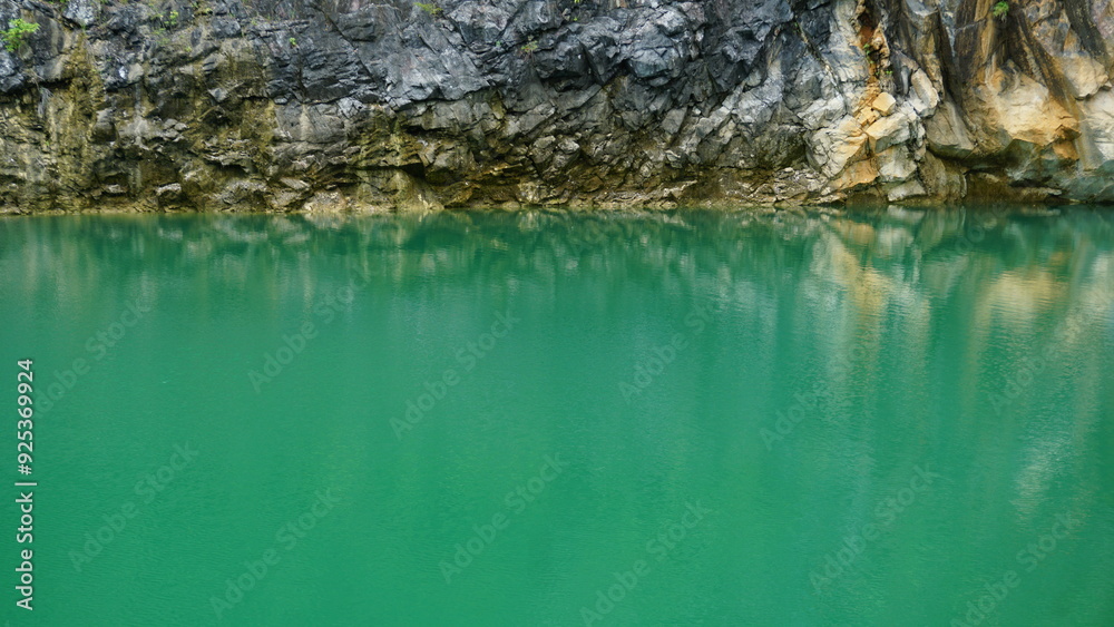 An image of a rocky cliff face reflected in the calm, emerald-green waters of a lake. An image of a rocky cliff face reflected in the calm, emerald-green waters of a lake.