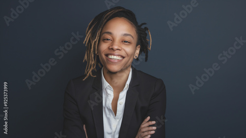 Nonbinary person with dreadlocks smiling against a dark background. They are wearing a black suit jacket and white shirt, conveying confidence and warmth. photo