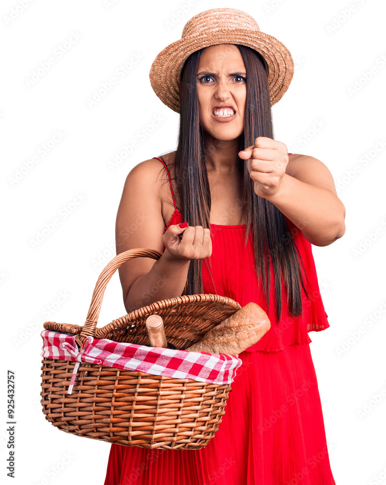 Young beautiful brunette woman wearing summer hat holding picnic basket annoyed and frustrated shouting with anger, yelling crazy with anger and hand raised Young beautiful brunette woman wearing summer hat holding picnic basket annoyed and frustrated shouting with anger, yelling crazy with anger and hand raised