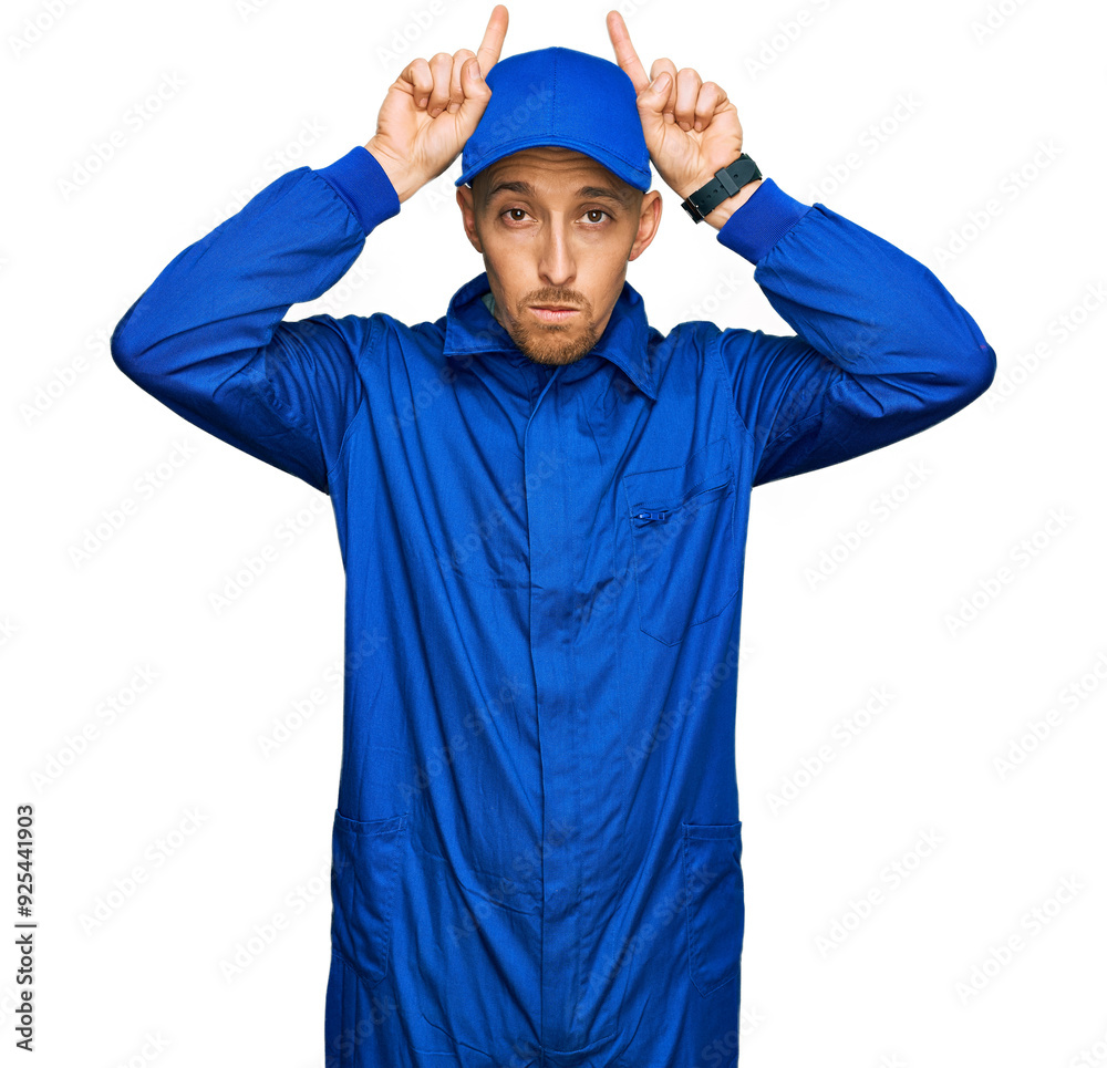 Bald man with beard wearing builder jumpsuit uniform doing funny gesture with finger over head as bull horns Bald man with beard wearing builder jumpsuit uniform doing funny gesture with finger over head as bull horns