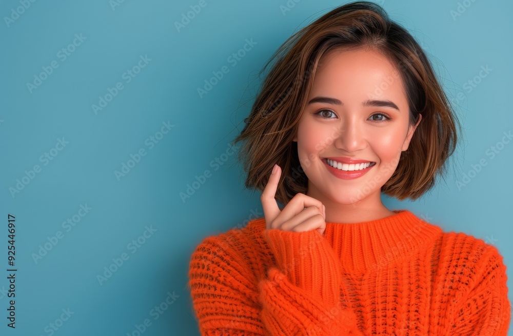 Young Woman Smiling While Pointing Up Against Blue Background in Cozy Orange Sweater Young Woman Smiling While Pointing Up Against Blue Background in Cozy Orange Sweater