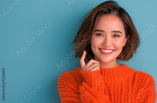 Young Woman Smiling While Pointing Up Against Blue Background in Cozy Orange ...