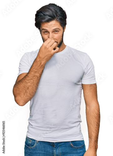 Young hispanic man wearing casual white t shirt smelling something stinky and disgusting, intolerable smell, holding breath with fingers on nose. bad smell