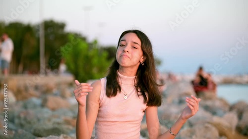 A young girl with dark skin is sitting on the rocks by the sea on a sunny summer day, looking at the camera and giving a thumbs up