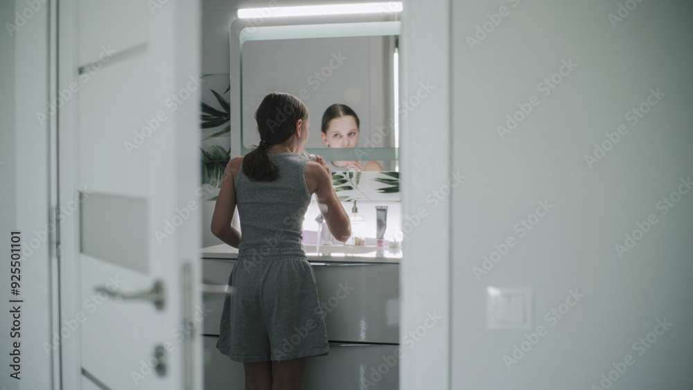 Young girl comes into bathroom, washes her face with cold water and looks at the mirror. Teenager stands in front of sink and uses electrical toothbrush to brush her teeth. Back view through door.