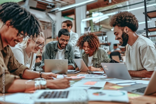 Smiling diverse team working together in a creative office environment, using laptops and papers to develop advertising ideas. High-resolution image showcasing teamwork and innovation.
