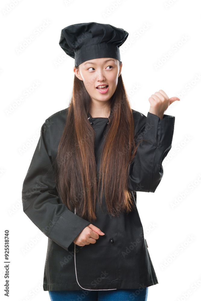 Young Chinese woman over isolated background wearing chef uniform smiling with happy face looking and pointing to the side with thumb up. Young Chinese woman over isolated background wearing chef uniform smiling with happy face looking and pointing to the side with thumb up.
