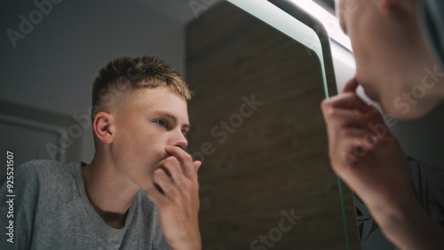 Caucasian boy stands in bathroom and looks at his reflection in the mirror. Teenager stands near wash basin and makes hairstyle in the morning. Bathroom routine concept. Lifestyle. Back view. Close up