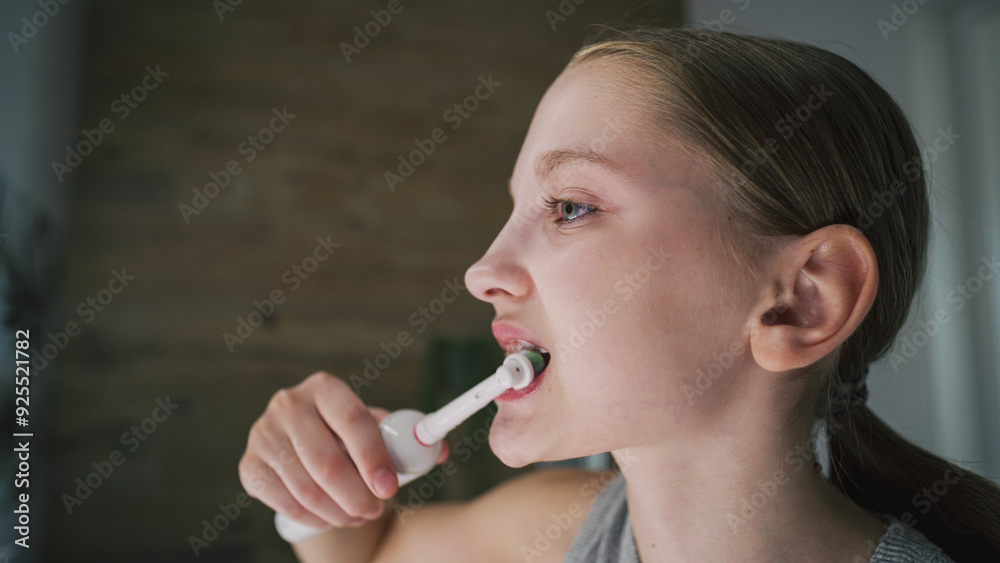 Young girl looks at mirror in bathroom, brushes her teeth. Pretty teenager stands in front of sink and uses electrical toothbrush before bedtime. Evening bath routine. Concept of lifestyle. Close up. Young girl looks at mirror in bathroom, brushes her teeth. Pretty teenager stands in front of sink and uses electrical toothbrush before bedtime. Evening bath routine. Concept of lifestyle. Close up.