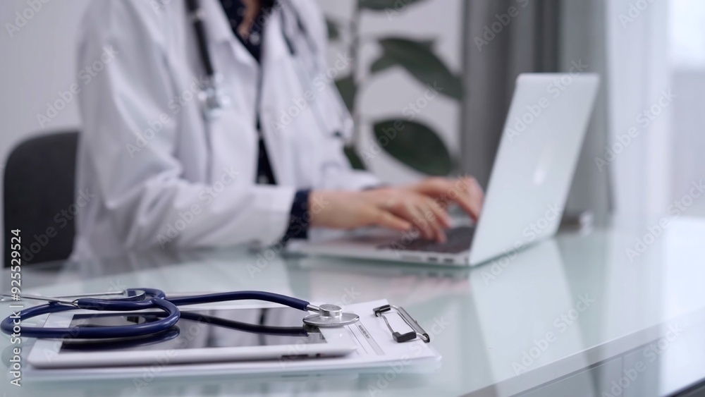 Tablet computer and blue stethoscope are on the glass table while doctor woman is using laptop computer on the glass table. Medicine concept Tablet computer and blue stethoscope are on the glass table while doctor woman is using laptop computer on the glass table. Medicine concept