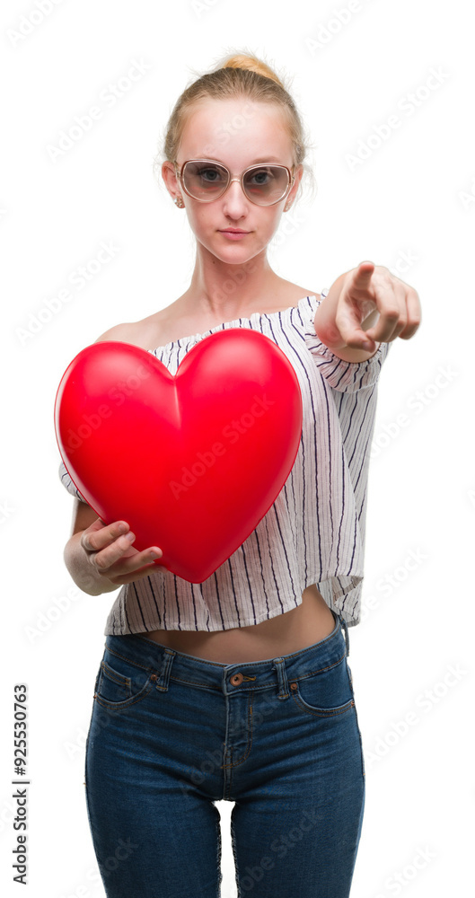 Blonde teenager woman holding red heart pointing with finger to the camera and to you, hand sign, positive and confident gesture from the front Blonde teenager woman holding red heart pointing with finger to the camera and to you, hand sign, positive and confident gesture from the front