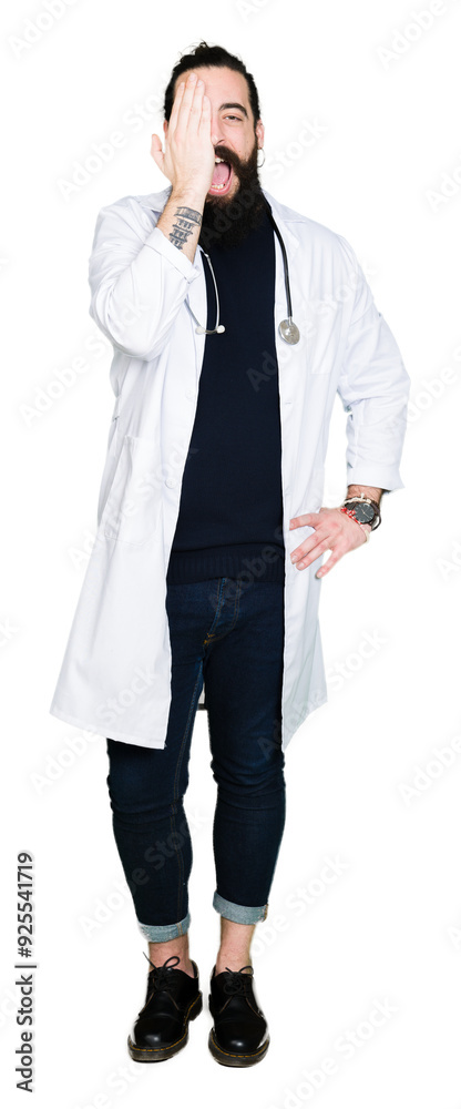 Doctor with long hair wearing medical coat and stethoscope covering one eye with hand with confident smile on face and surprise emotion. Doctor with long hair wearing medical coat and stethoscope covering one eye with hand with confident smile on face and surprise emotion.