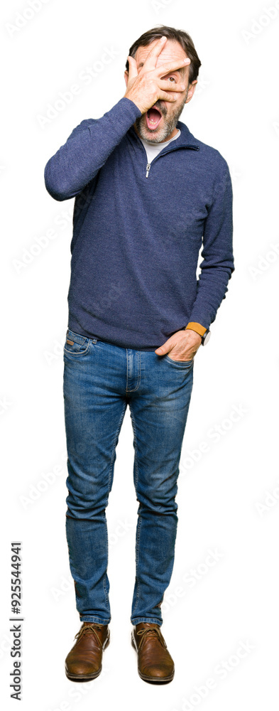 Middle age handsome man wearing a sweater peeking in shock covering face and eyes with hand, looking through fingers with embarrassed expression. Middle age handsome man wearing a sweater peeking in shock covering face and eyes with hand, looking through fingers with embarrassed expression.