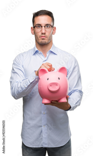 Handsome young man holding a piggy bank with a confident expression on smart face thinking serious