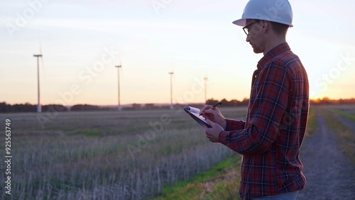 Man engineer wearing a white protective helmet is taking notes with a clipboard in a field with wind turbines, as the sun sets. Clean energy and engineering concept