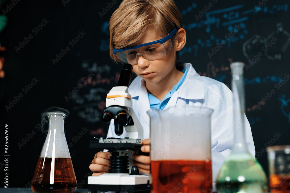 Blonde hair schoolboy in laboratory wear lab coat stand and learn science of chemistry technology in STEM class. The student looking object in microscope . On the table put many flask. Erudition. Blonde hair schoolboy in laboratory wear lab coat stand and learn science of chemistry technology in STEM class. The student looking object in microscope . On the table put many flask. Erudition.