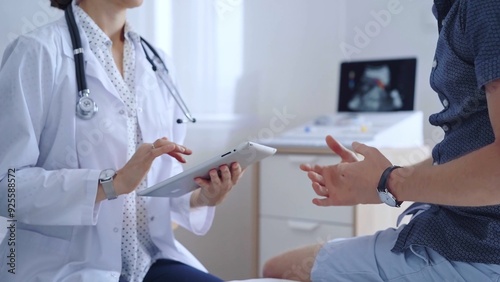 Doctor woman is taking notes on a tablet computer while a male patient describes his symptoms in clinic procedure cabinet. Medicine service concept