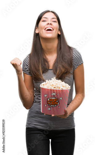 Young beautiful hispanic eating popcorn screaming proud and celebrating victory and success very excited, cheering emotion