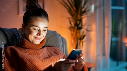 Smiling woman relaxes on a cozy chair, managing her smart home system effortlessly through her smartphone, showcasing seamless technology integration.