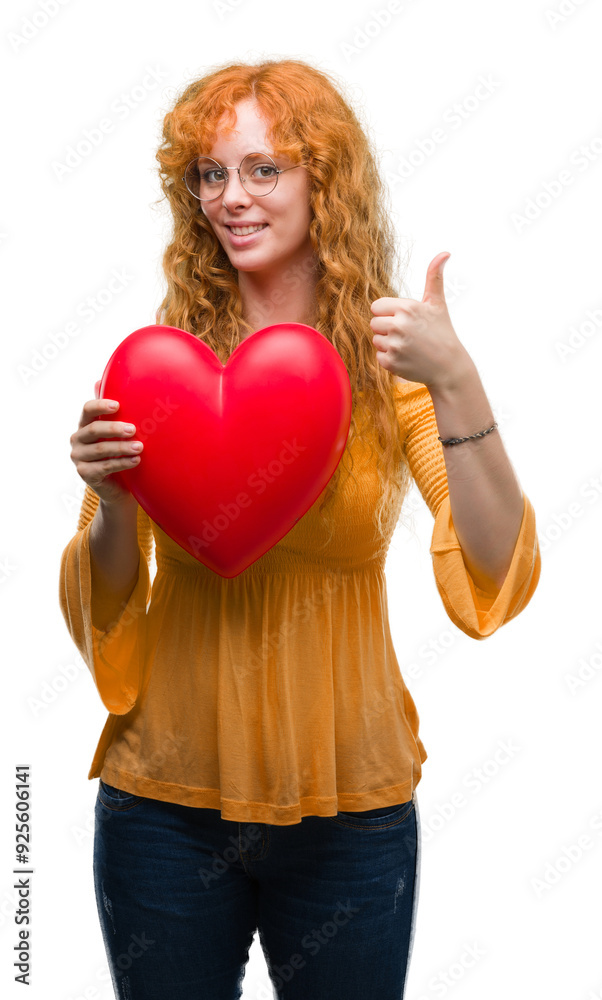 Young redhead woman in love holding red heart happy with big smile doing ok sign, thumb up with fingers, excellent sign Young redhead woman in love holding red heart happy with big smile doing ok sign, thumb up with fingers, excellent sign