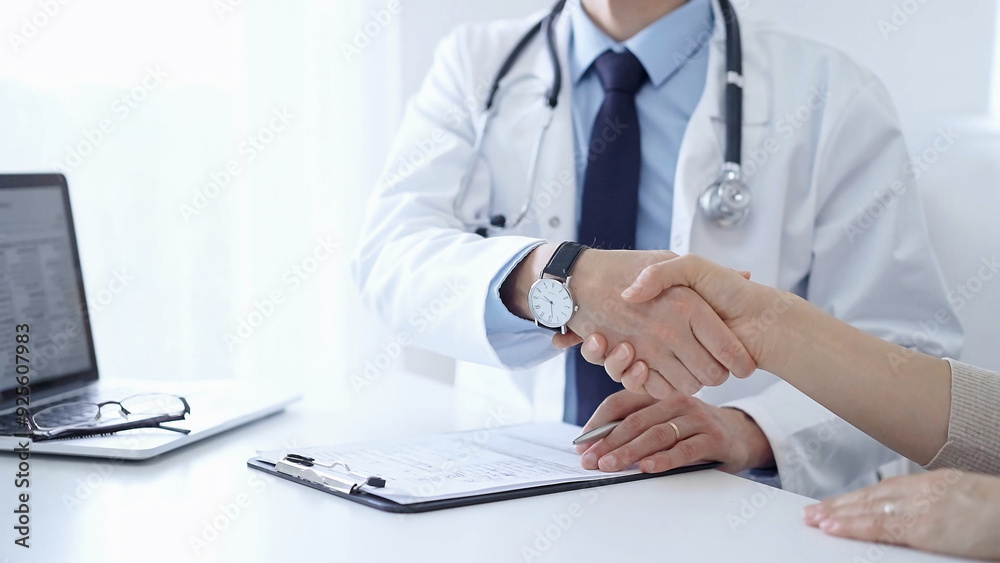 Doctor and a patient are shaking hands. The physician is wearing a white medical coat over a blue shirt. Medical service Doctor and a patient are shaking hands. The physician is wearing a white medical coat over a blue shirt. Medical service