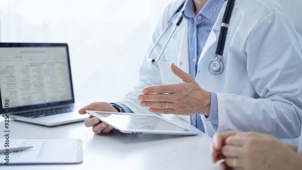 Doctor and a patient. The physician, wearing a white medical coat over a blue shirt, is using tablet computer during a consultation in the clinic. Medicine and science Doctor and a patient. The physician, wearing a white medical coat over a blue shirt, is using tablet computer during a consultation in the clinic. Medicine and science