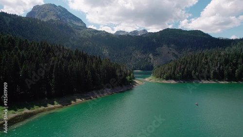 A mountain lake below a mountain peak surrounded by a coniferous forest at dawn captured by a drone in motion.