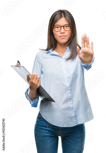 Young asian business woman over isolated background holding clipboard with open hand doing stop sign with serious and confident expression, defense gesture