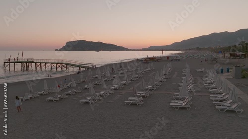 A serene beach scene at sunset, featuring rows of empty loungers facing the calm sea, with a pier extending into the water and mountains in the background. photo