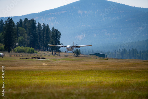 Twin Otter airplane takes off over field and in front of pine tree forest and a mountain