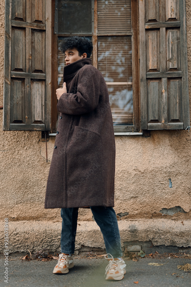 Fashionable man in a brown coat and jeans standing in front of an old building with shuttered windows