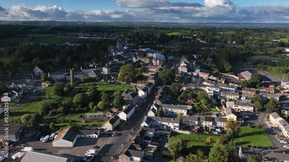 Clones Town, County Monaghan, Ireland, April 2023. Drone descends orbiting clockwise in a panoramic aerial establishing town with Round Tower surrounded by buildings and cemetery on a cloudy day. Clones Town, County Monaghan, Ireland, April 2023. Drone descends orbiting clockwise in a panoramic aerial establishing town with Round Tower surrounded by buildings and cemetery on a cloudy day.