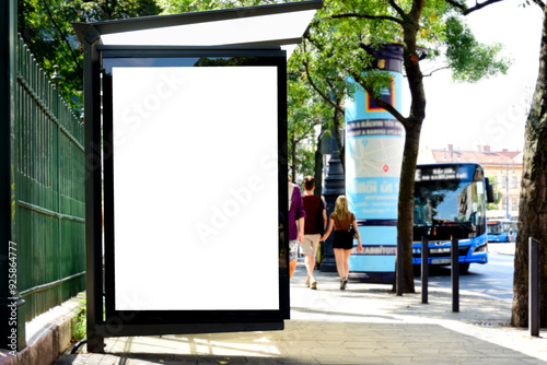 Fototapeta Naklejka Na Ścianę i Meble -  bus shelter at busstop. white poster ad display space and lightbox. mockup template. blank ad panel. glass design. urban street setting. green background. silhouettes in the background