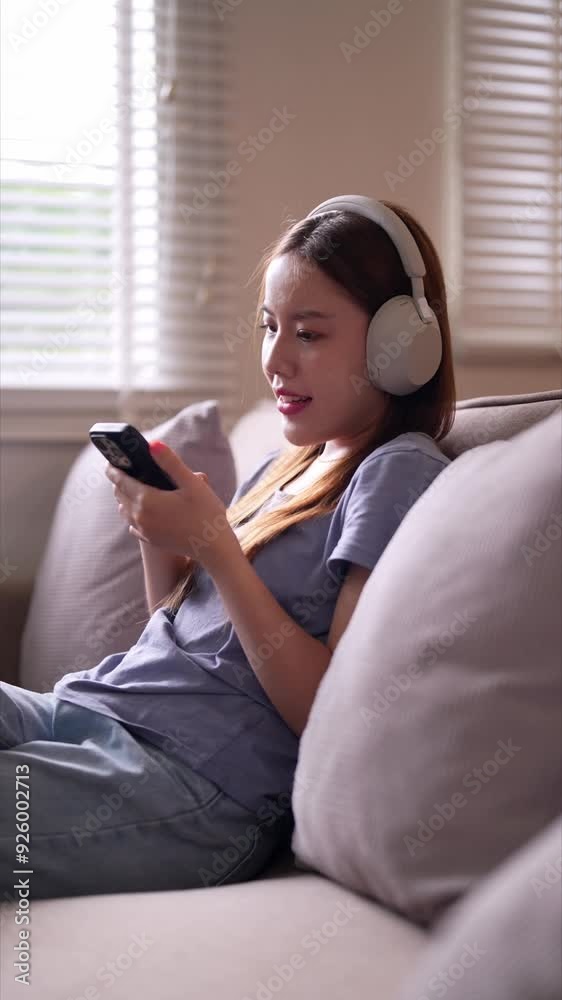 A young woman sitting on a sofa, wearing headphones, smiling while looking at her phone, enjoying music or a video in a cozy home environment A young woman sitting on a sofa, wearing headphones, smiling while looking at her phone, enjoying music or a video in a cozy home environment