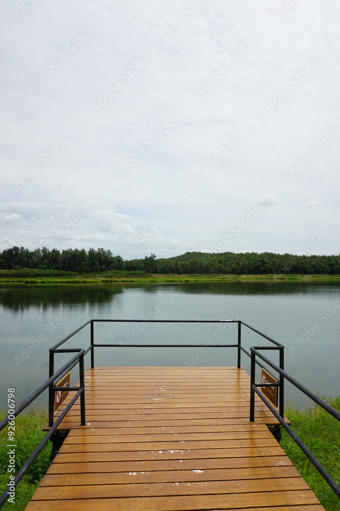 Obraz premium A tranquil scene of a wooden pier extending into a calm lake. The pier has a metal railing and offers a peaceful view of the surrounding natural scenery. Lush green hills and a cloudy sky complete the