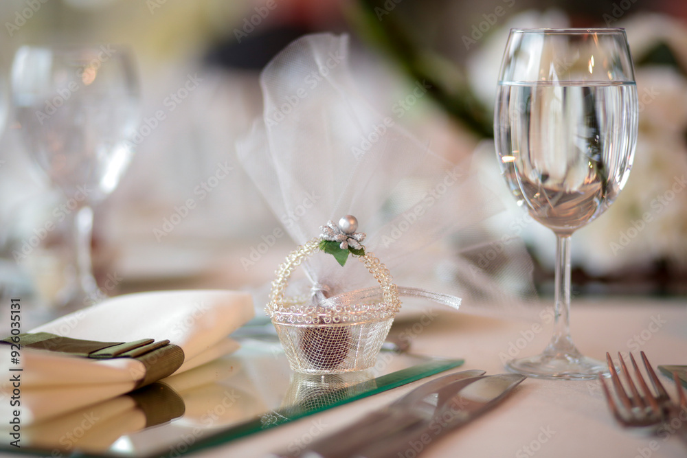 A close-up of a wedding favor on a beautifully decorated table setting. The favor is a small box wrapped in tulle and tied with a ribbon
