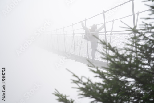 Fearless woman crossing a suspension bridge. Safety rope. Tourist suspension bridge. Concept of adrenaline, risk, challenge and obstacle. Courage walk over abyss- Foggy day photo