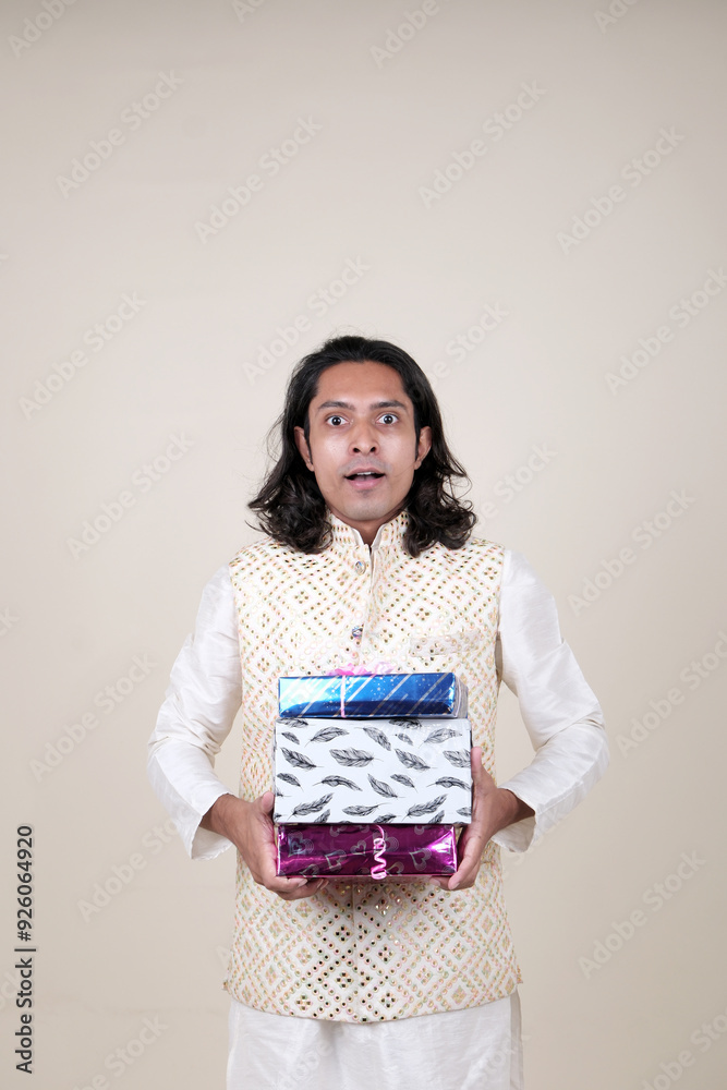 A happy young man in traditional attire, holding gift boxes and looking at the camera against a cream background. A happy young man in traditional attire, holding gift boxes and looking at the camera against a cream background.