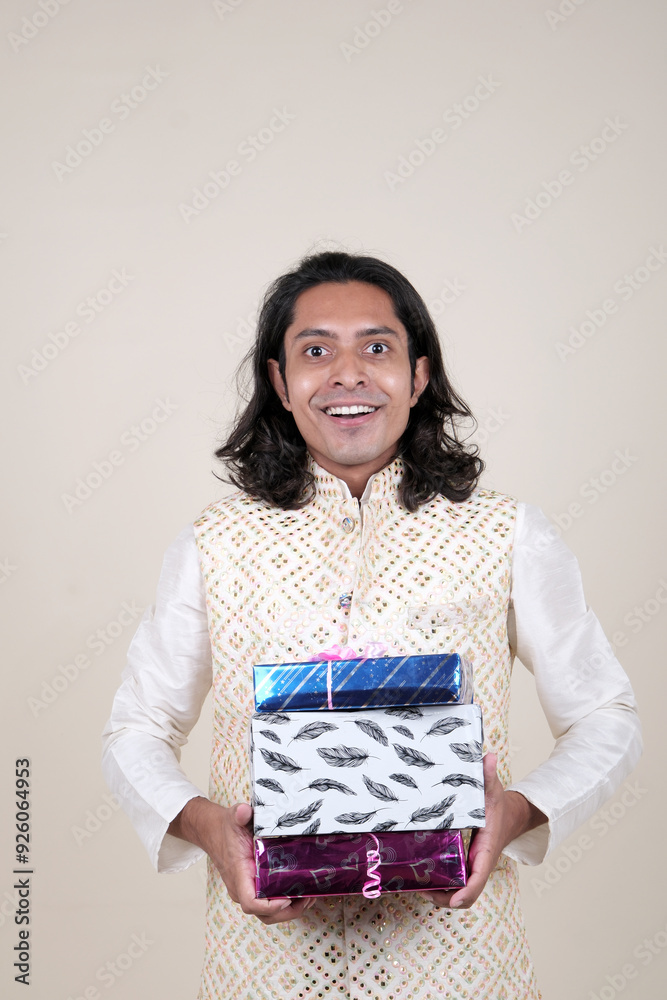 A happy young man in traditional attire, holding gift boxes and looking at the camera against a cream background. A happy young man in traditional attire, holding gift boxes and looking at the camera against a cream background.